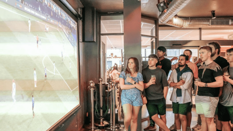 A huge HD screen in a Belushi's Bar with football fans watching the game gathered round.