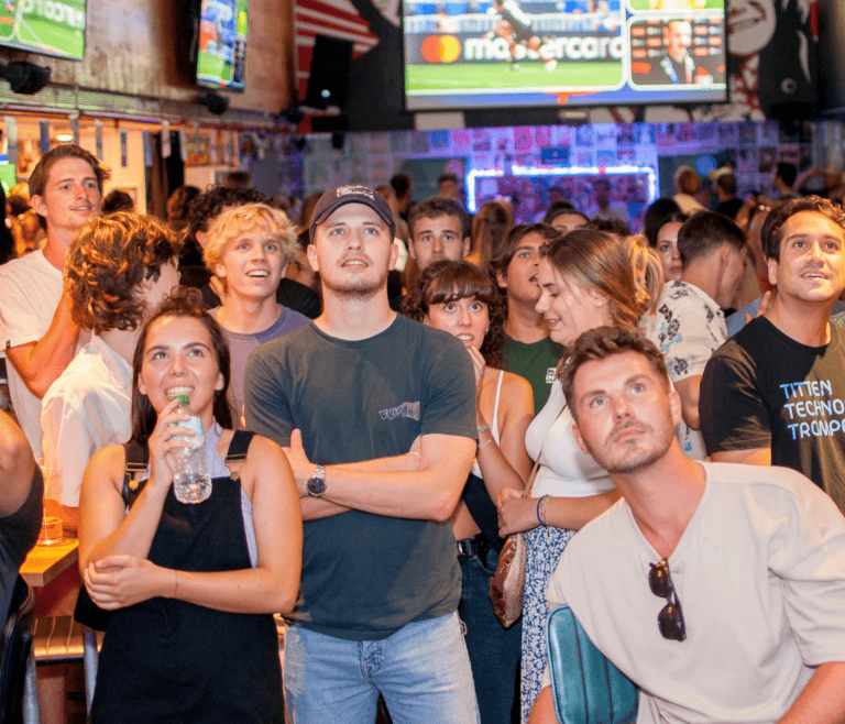 Customers at a Belushi's bar standing and watching large TV screens expectantly