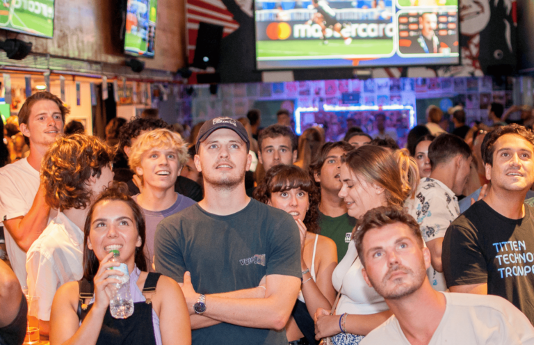 Customers at a Belushi's bar standing and watching large TV screens expectantly