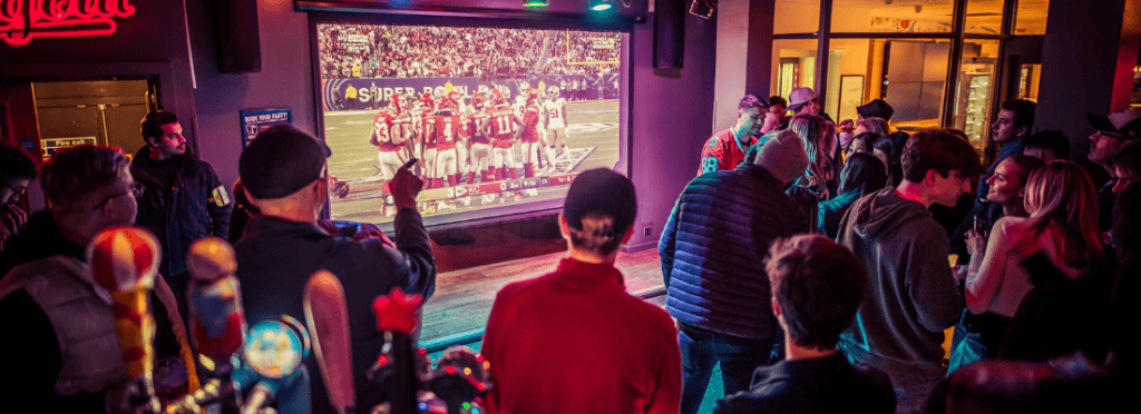 A large group of fans watches Super Bowl on a huge projector n a Belushi's Bar.