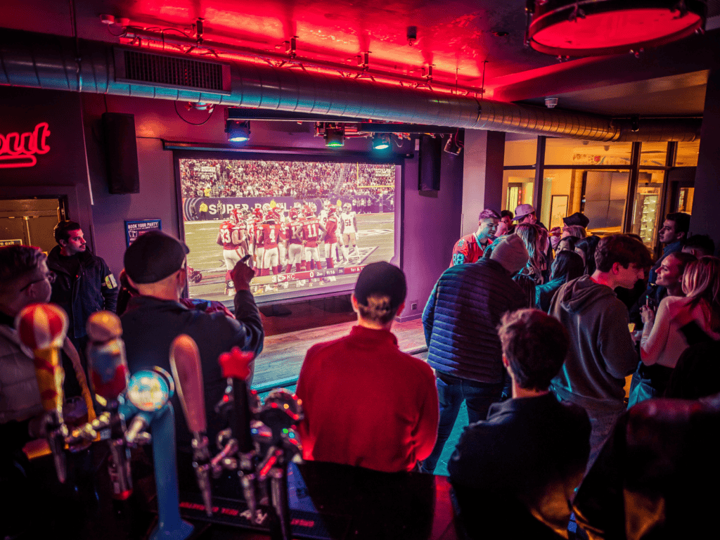 A large group of fans watches Super Bowl on a huge projector n a Belushi's Bar.