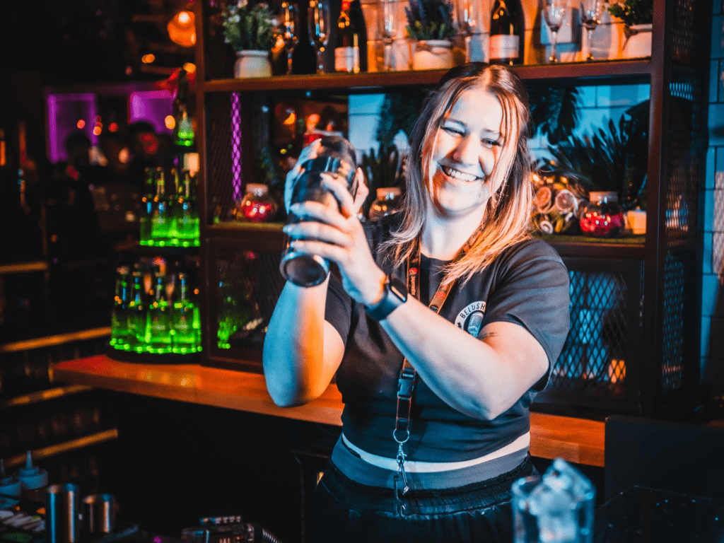 A friendly bartender smiles and shakes a cocktail at a Belushi's Bar.