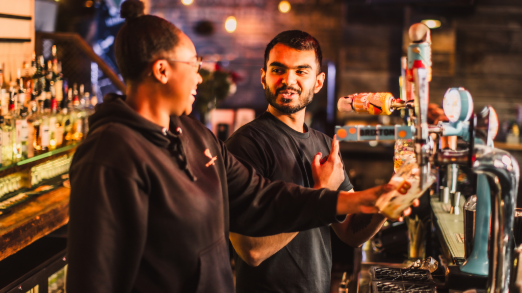 Two bartenders smiling and laughing while pouring pints at Belushi's London Bridge bar.