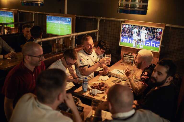 An image of a group sat at a booth watching football while sharing wings and drinking pints