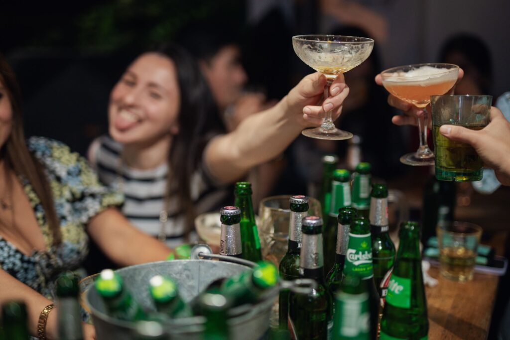 An image of people at a table, cheersing with a beer and a few cocktails, as well as buckets of beers on the table