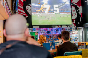 An image of a large HD screen with seated customers watching rugby at a Belushi's bar.