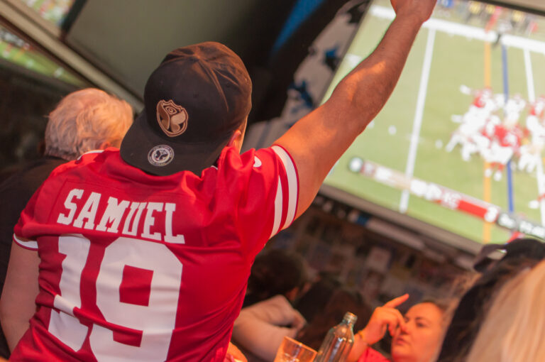 An image of a man, from behind, raising his arm in celebration of a game on a large projector in the background.