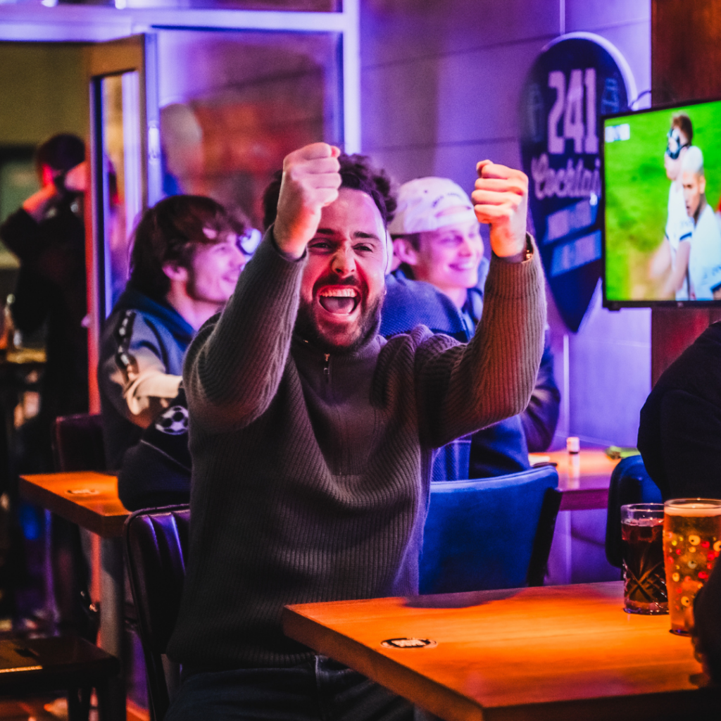 A person celebrating a Premier League goal at a Belushi's bar, with drinks on the table.