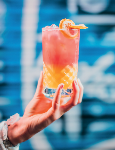 A hand holding out a pink and orange cocktail with a blue background.