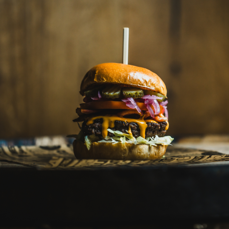 A burger with a darkened background, with pickles, tomato, onion, cheese and lettuce.