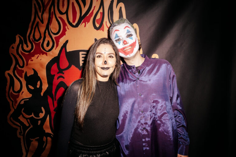 A couple in Halloween makeup posing together: woman with skull-face makeup and black outfit, man with clown makeup and purple shirt against a patterned backdrop.