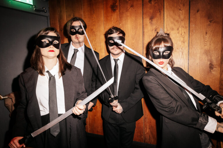 Four people in black suits and masks pose with swords against a wooden wall, as if at a costume or performance event.