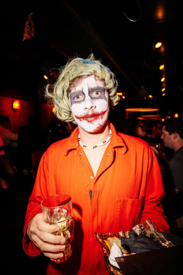 Person wearing orange jumpsuit with Joker makeup at a party, holding a glass of drink in one hand and a tray of snacks in the other.