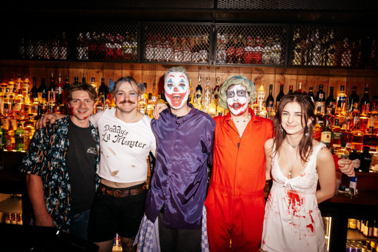 Five friends in Halloween costumes pose at a bar, including two clowns and a blood-splattered maid, smiling with liquor shelves in the background.