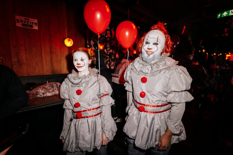 Two clowns in gray costumes with red pom-poms and white face paint walk through a dim, red-lit party scene with balloons nearby.