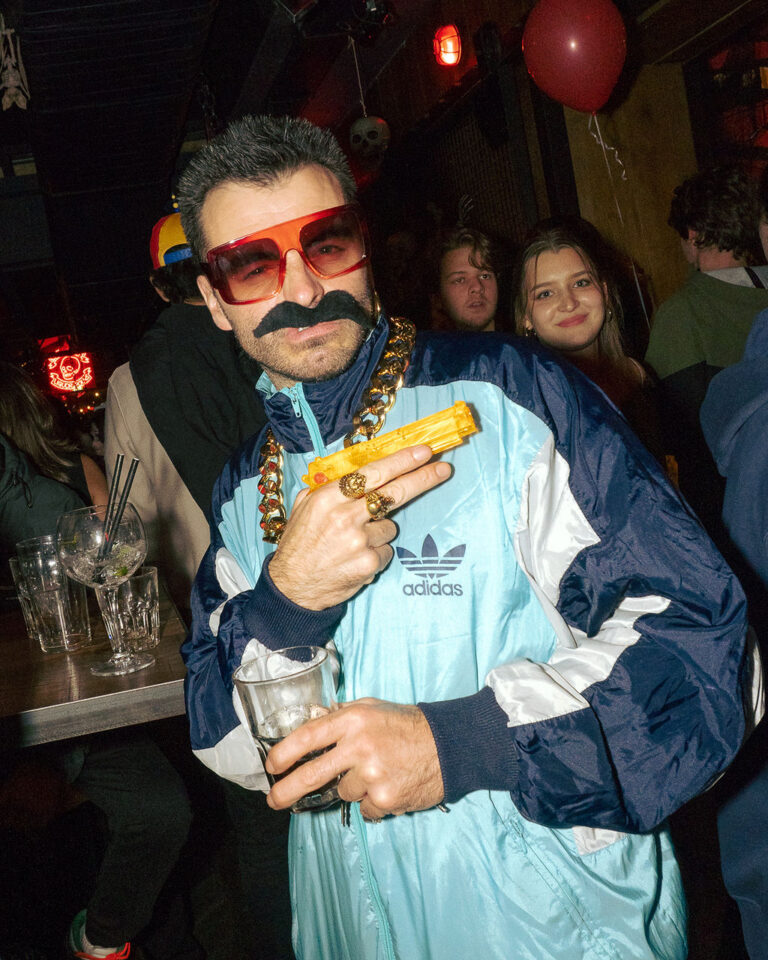 Man at a party wearing oversized red sunglasses and a fake mustache, holding a yellow toy gun and a drink, with a gold chain and blue Adidas jacket.
