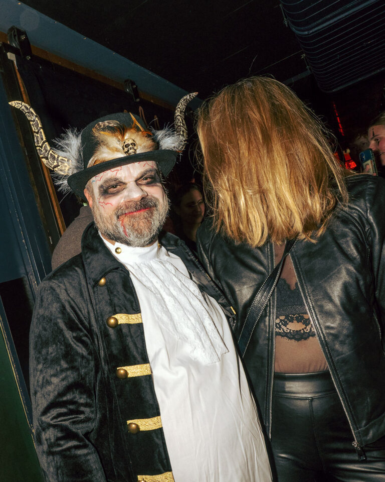 Man in Halloween makeup with skull-horned hat and velvet coat smiles at the camera at a party.