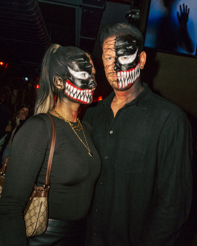 Couple at a nightclub with scary skull makeup, posing for the camera; woman in black top with a gold necklace and a Gucci handbag, man in a black shirt.