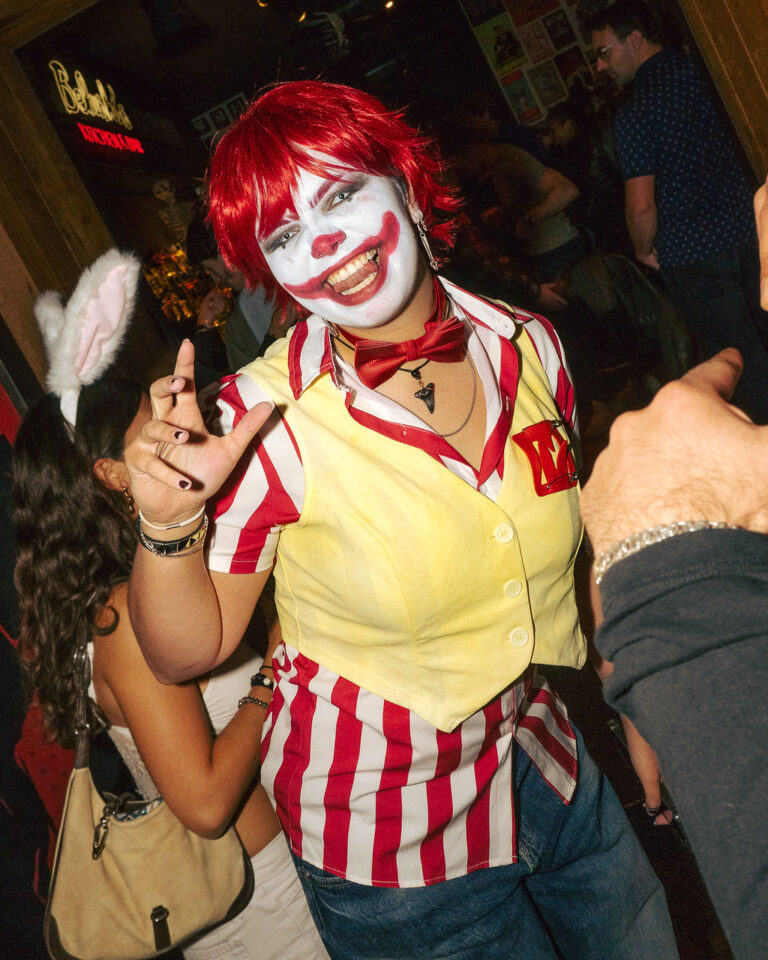 Person in clown costume with red wig, white face paint, red nose, and red lipstick posing with fingers extended at a crowded party.