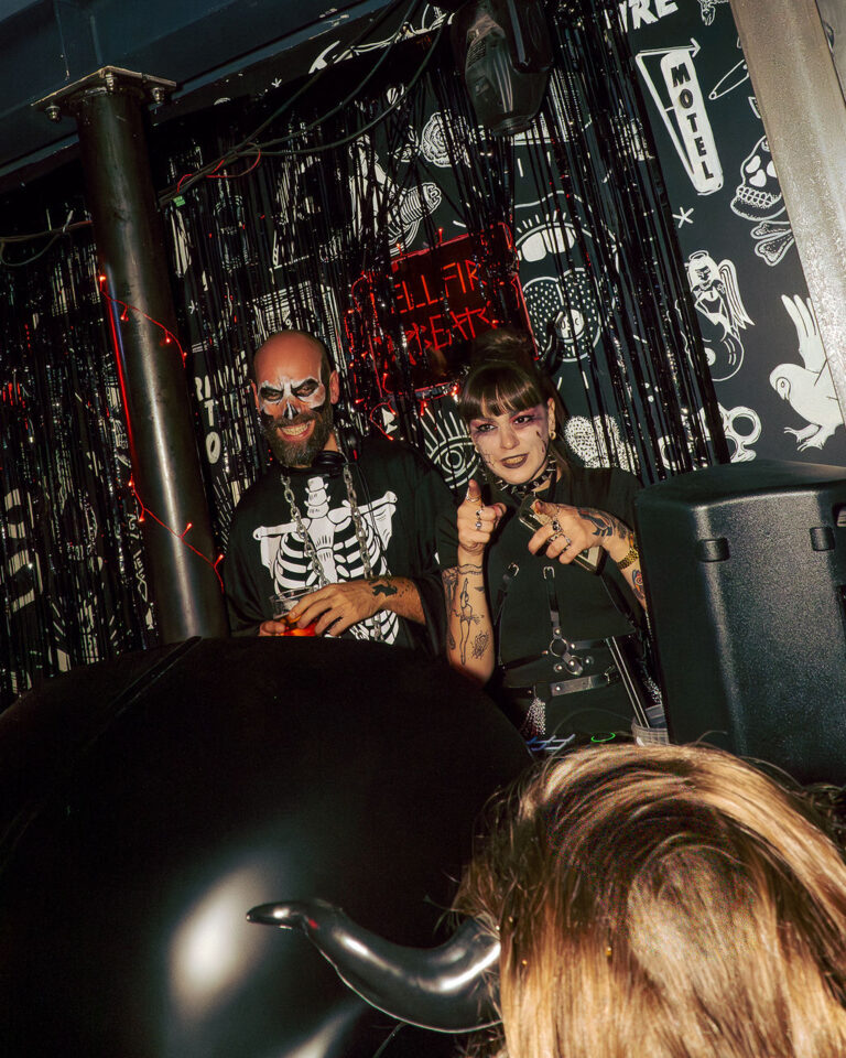 Two people in Halloween outfits pose behind a DJ booth in a dark club; skeleton-shirted man with face paint, woman with dramatic makeup pointing at the camera.