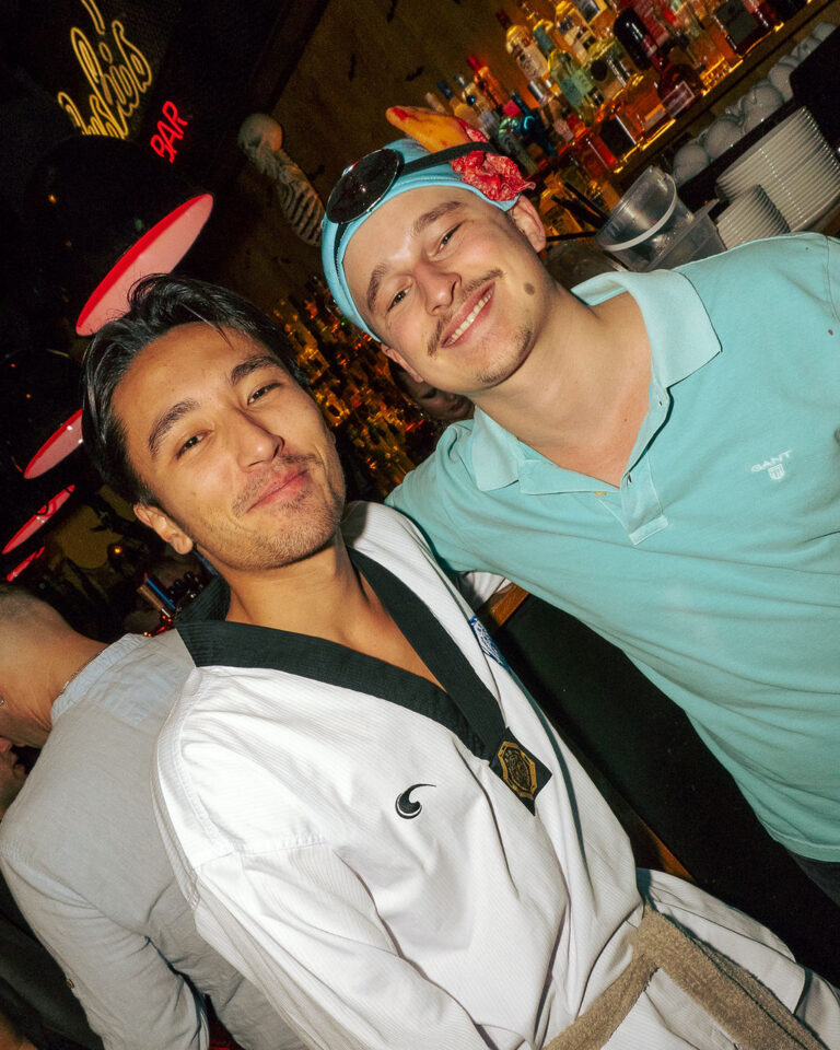 Two men smiling at the camera in a bar; one wears a white martial arts gi with a black belt and medal, the other wears a light blue polo and a headband with sunglasses on his head.
