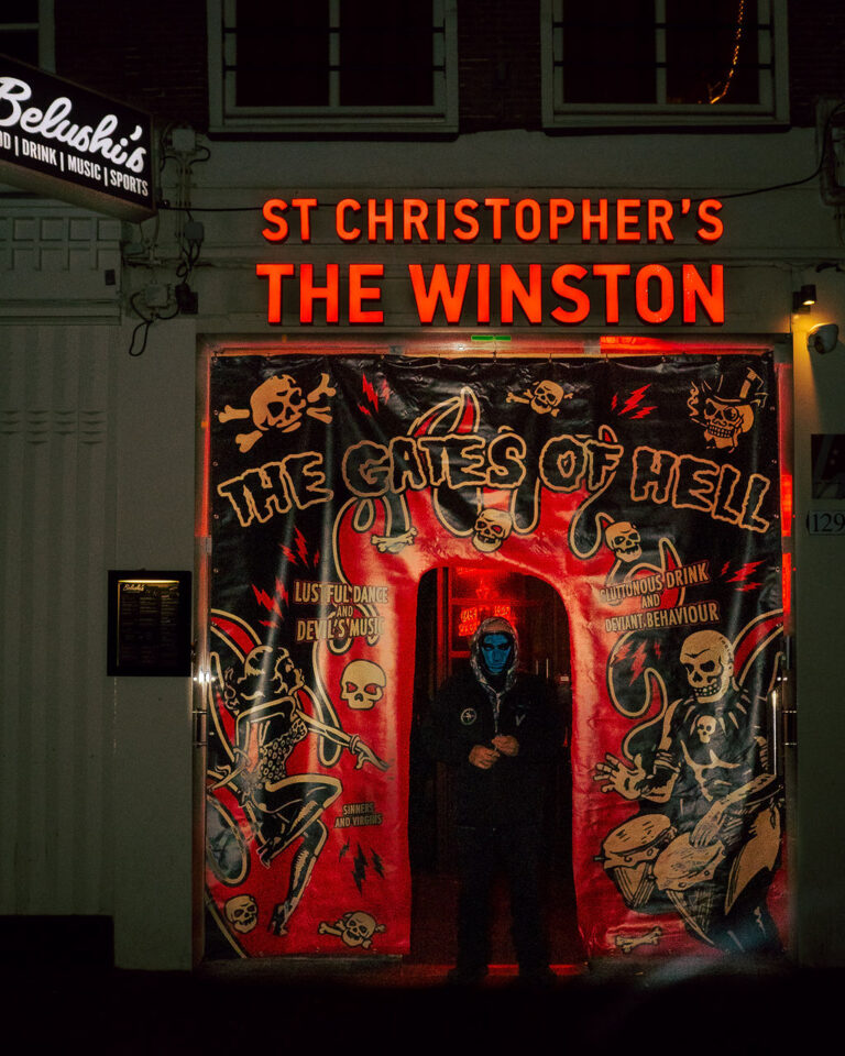 Night club entrance at night with red neon sign 'ST CHRISTOPHER'S THE WINSTON' and a person in a blue mask/hood standing in a red-lit doorway with skull artwork above