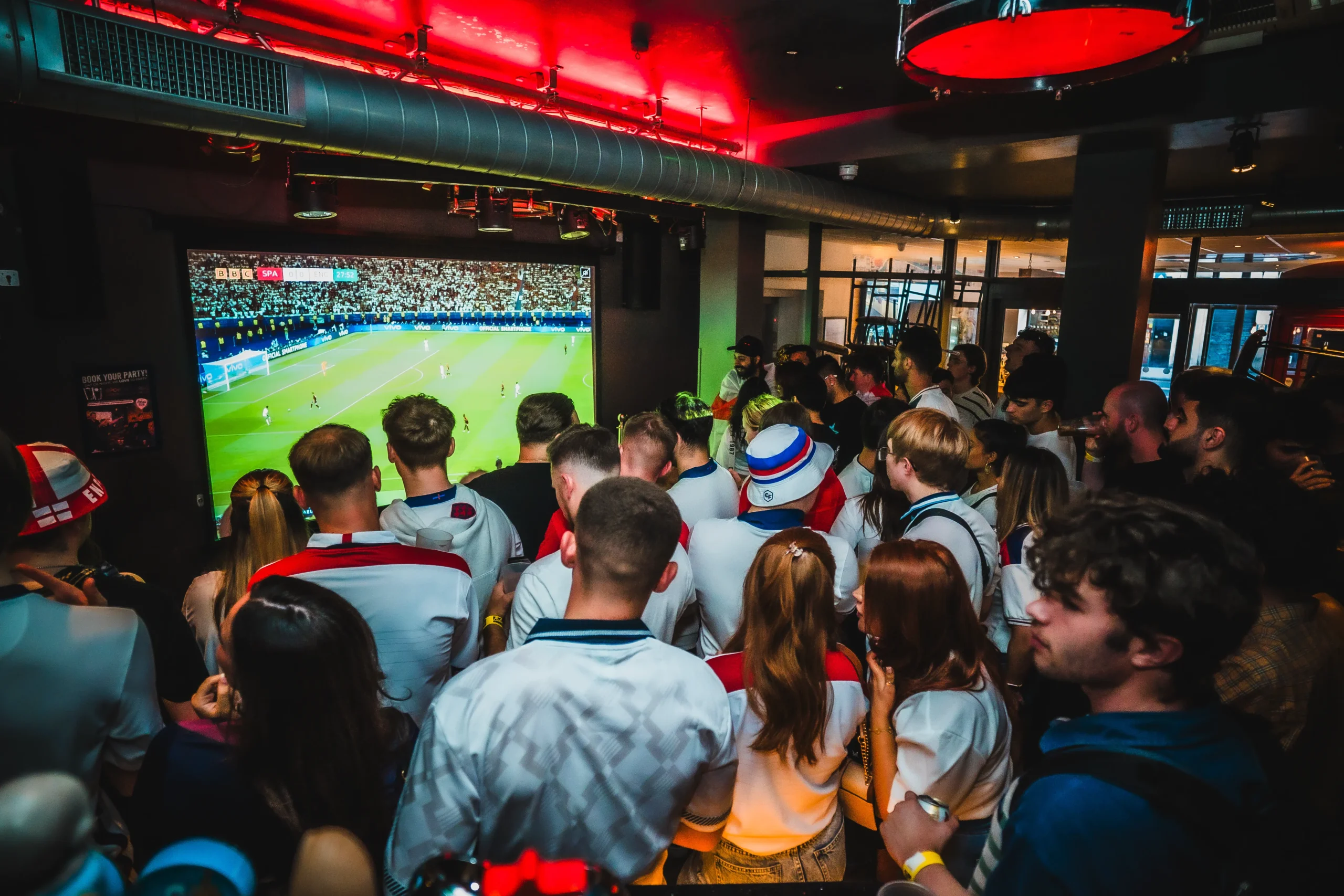 Crowd of football fans watching a World Cup 2026 match live on a large projector screen at Belushi's Greenwich, with red overhead lighting and a packed standing crowd