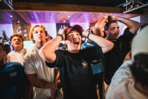 Crowd of excited young people with raised arms at a lively indoor event; front center person wears a red cap and screams with joy.