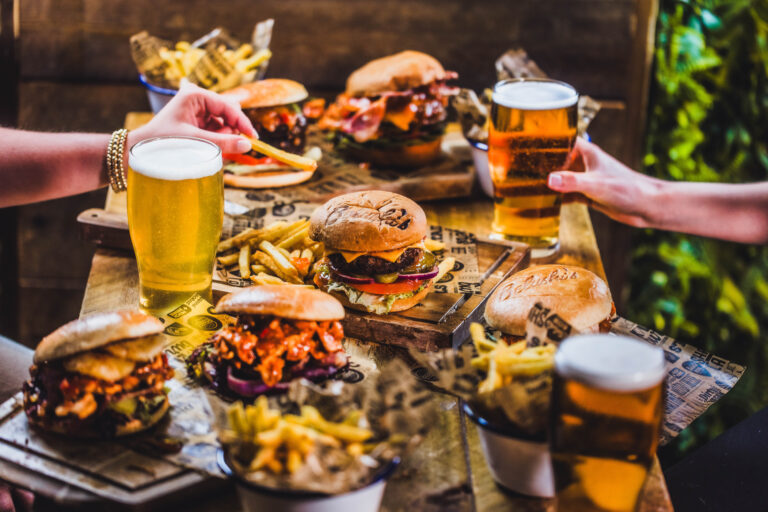 Two people clinking pint glasses over a wooden table full of burgers, fries, and condiments