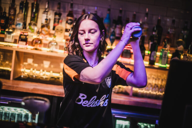 Female bartender shaking a cocktail behind a bar illuminated with colorful lights.