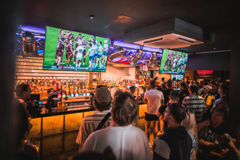 Bar interior with multiple TVs showing a rugby match, crowded with patrons at the counter and bottles visible behind them.