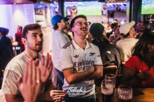 Group of friends at a busy bar watching a sports game, central man in a white Arsenal/Airways shirt smiling and arms crossed while others clap nearby.