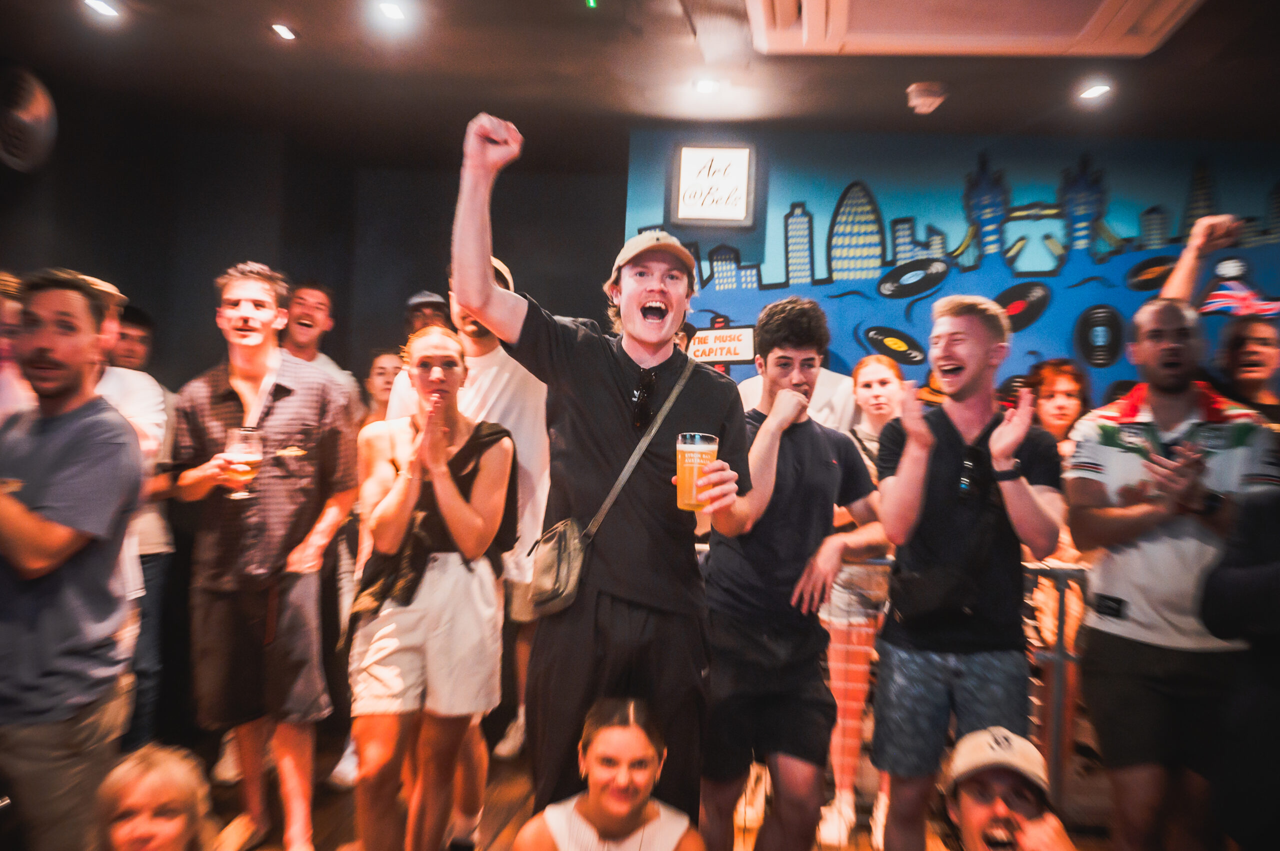 Crowd of people at a party cheering, central man raises a fist and holds a beer with a bright mural behind them.