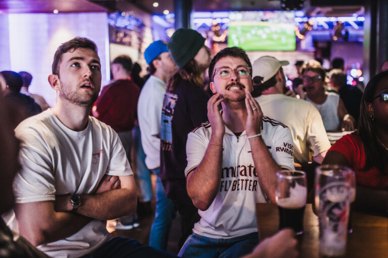Group of friends at a dim sports bar watching a game on a TV screen in the background, one man adjusting his mustache while glasses-wearing man looks up in anticipation, beer glasses on the table nearby.