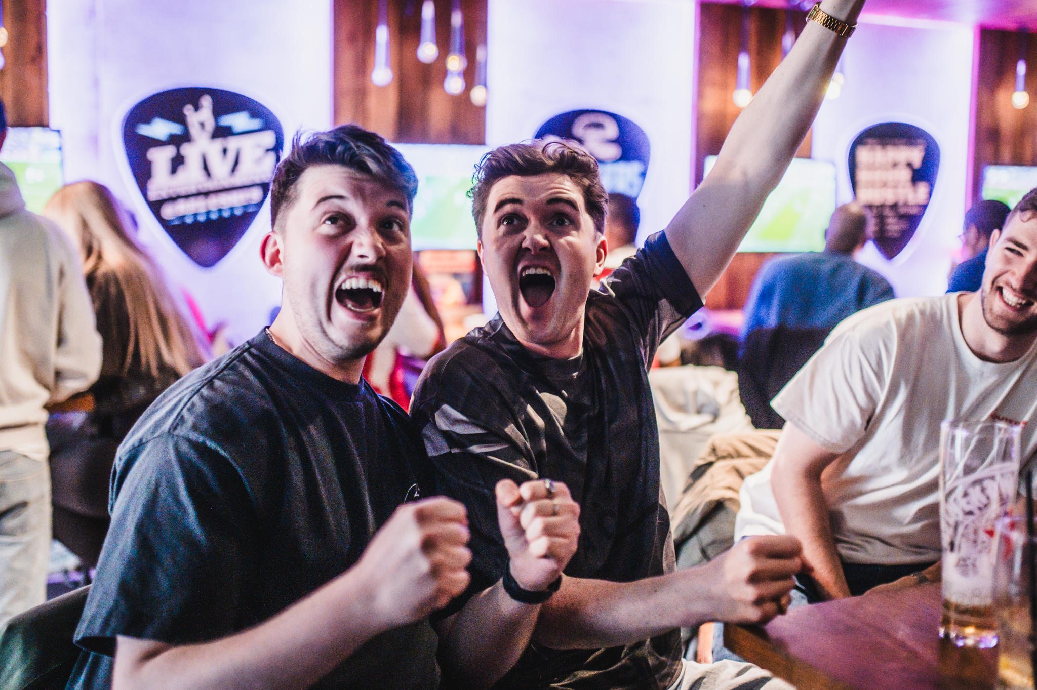 Three friends at a lively bar cheering with raised fists toward the camera, neon lights in the background.