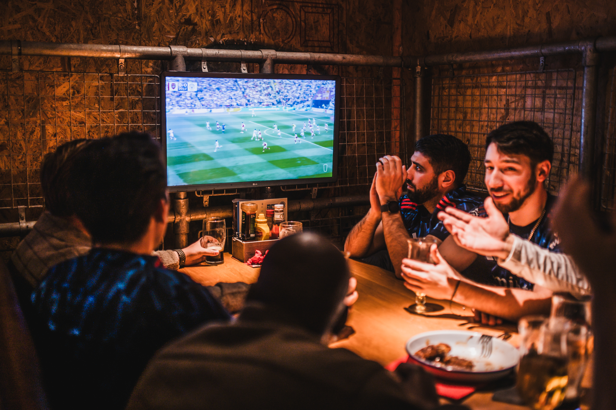 Group of friends at a dim, industrial bar watching a soccer match on a wall-mounted TV with drinks on the table.