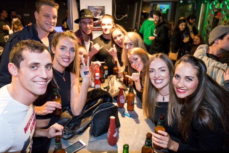 Group of young friends at a party posing for a photo around a table with beer bottles, smiling for the camera (indoors).