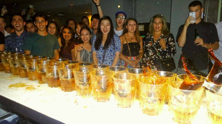 Group of young adults at a party standing behind a bar lined with dozens of orange shot glasses filled with drink, some splashing.
