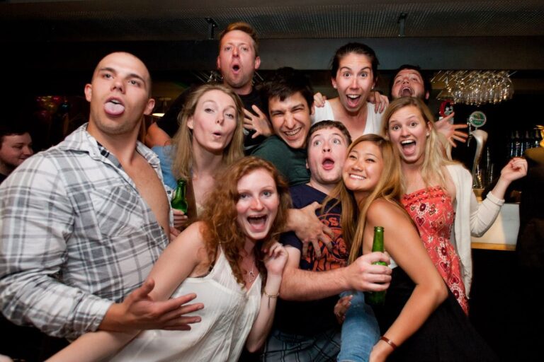 A group of friends at a party posing closely for a photo, some sticking out their tongues and cheering, holding drinks and smiling at the camera.