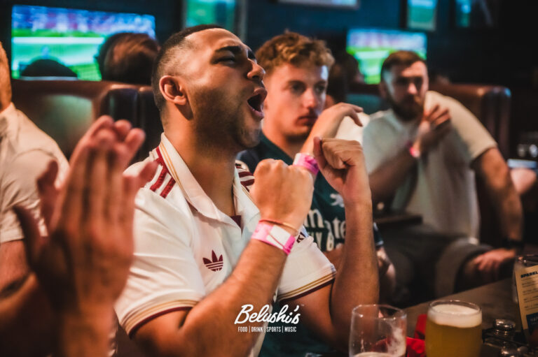Man in a white Adidas shirt cheers with raised fists as friends watch in a lively bar scene at Belushi's.