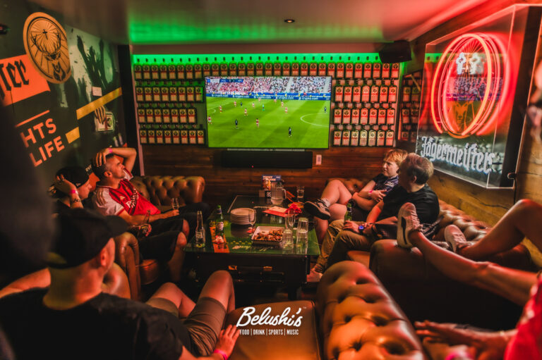 Group of friends watching a soccer match on a large TV in a neon-lit sports bar with bottles lining the back wall for decoration.