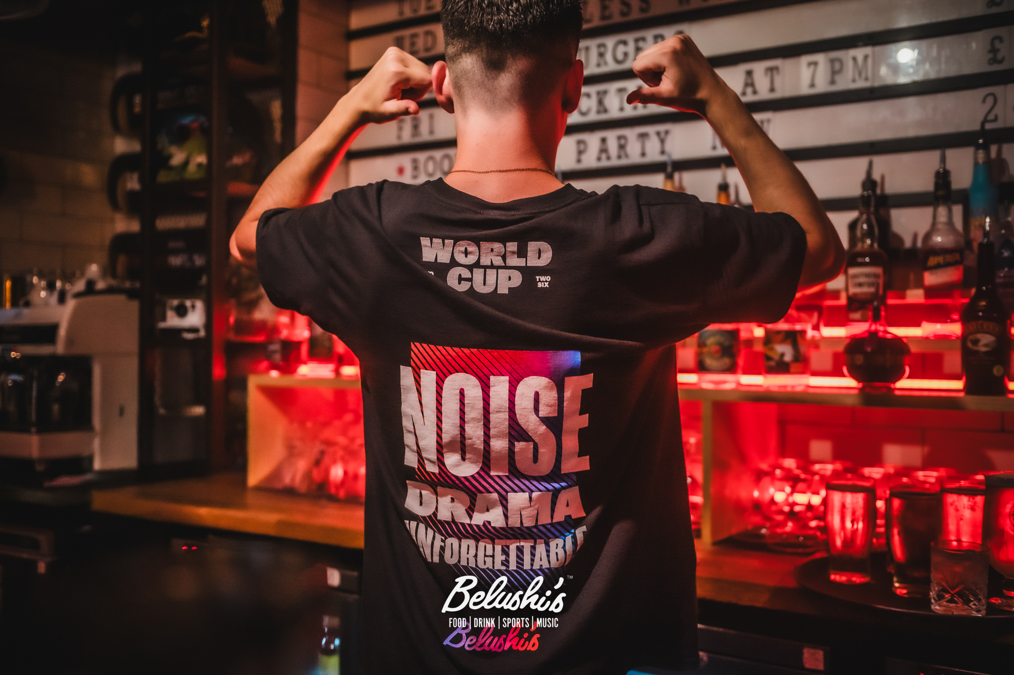 Back view of a man in a dark t-shirt facing a bar, arms raised, in a red-lit bar interior (Belushi's).