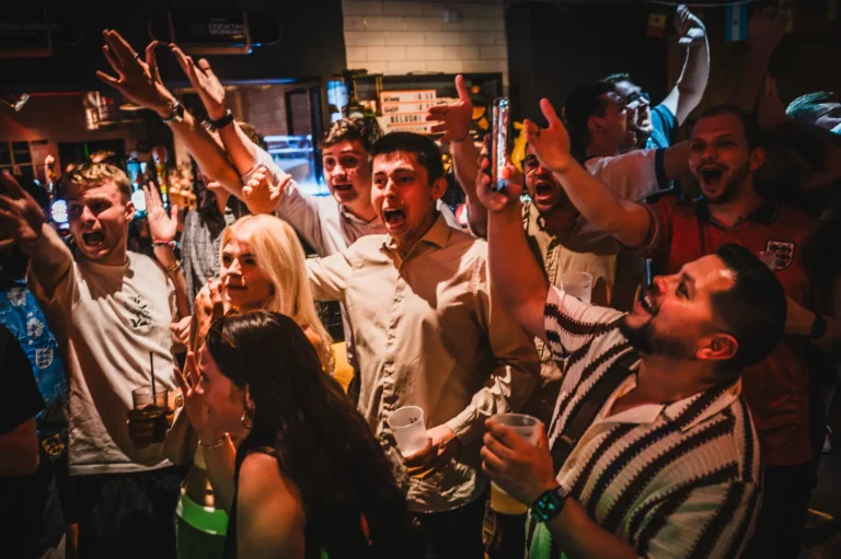 Crowded party scene with people raising hands, cheering, and holding drinks in a dimly lit bar.