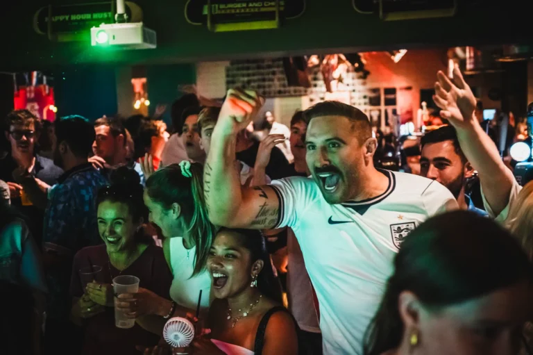 A crowded party scene with people cheering and smiling, one man in a white England soccer jersey raises his arm in celebration while others hold drinks at a dim, colorful venue.