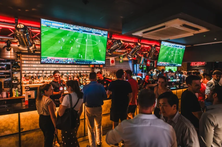 Crowded sports bar with several TVs showing a soccer match and neon red lighting behind the bar.
