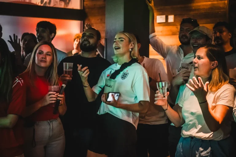 A group of friends at a crowded party, holding drinks and cheering toward something off-camera.