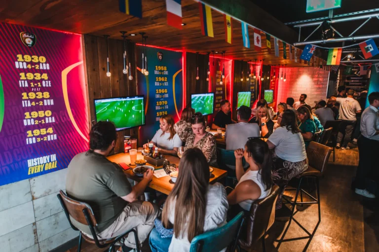 Crowded sports bar with people watching a soccer match on wall TVs and colorful wall panels above. Flags hang from the ceiling as warm lighting glows over the tables.