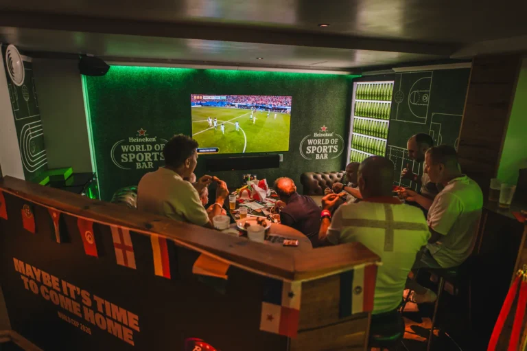 Group of people watching a soccer match on a large screen in a Heineken World of Sports Bar, with drinks and food on the table.