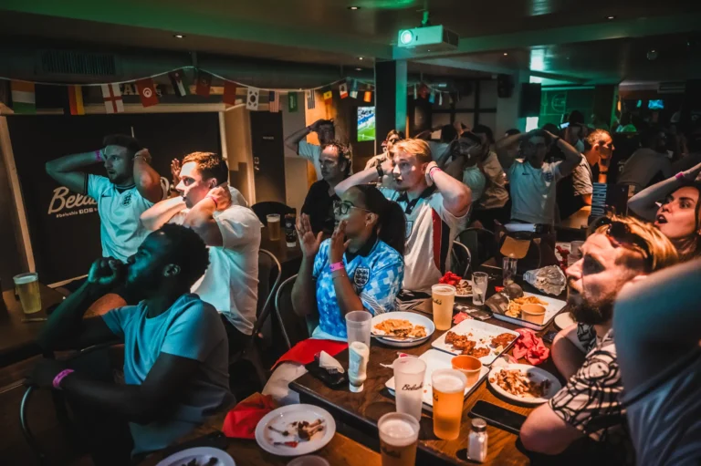 Crowded bar with soccer fans reacting to a game, some holding their heads, beer and plates of food on the table in front of them.
